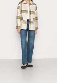 Rutig mönstrad jacka i beige och olivgrön, med knäppt front, bröstficka och avslappnad passform, tillsammans med blå raka jeans.