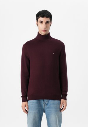 Maglione a collo alto color borgogna in morbido tessuto maglia con polsini e orlo a costine. Piccolo logo in evidenza sul petto. Abbinato a jeans azzurri chiari.