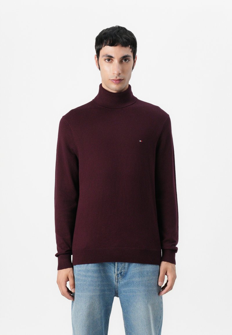 Pull à col roulé bordeaux en doux tricot, avec des poignets et un ourlet côtelés. Petit logo sur la poitrine. Assorti à un jean bleu clair.