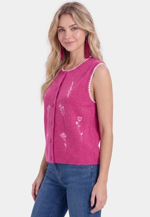 Kvinde med langt blond hår iført magentafarvet ærmeløs cardigan med hvid blomsterbroderi og blå jeans, som smiler let, på hvid baggrund.