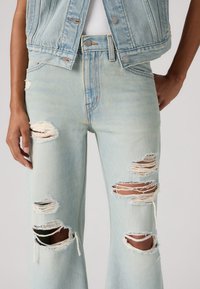 Jean bleu clair déchiré avec de grands trous effilochés aux cuisses et aux genoux, porté avec une veste en jean courte et un haut blanc.