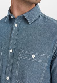 Camicia di jeans in blu sbiadito con colletto a punta, bottoni bianchi e cuciture in contrasto bianche. Presenta due tasche sul petto.