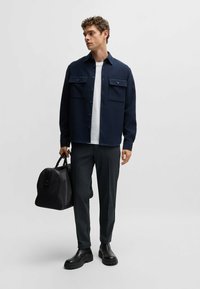 Navy button-up shirt with two front pockets, white t-shirt underneath, gray trousers, black leather boots, and a black bag.