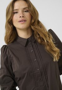Chemise marron foncé à manches longues avec un col boutonné, un devant plissé et des manches bouffantes. Elle présente des boutons assortis au tissu.