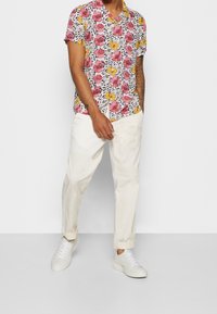 Korte mouwen, knoopsluiting overhemd met een kleurrijk bloemenpatroon; gedragen met crèmekleurige, taps toelopende broek en witte sneakers.