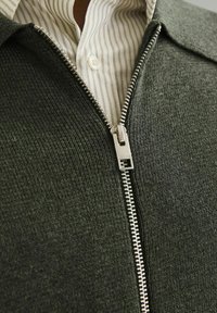 Sivi zip-up džemper izrađen od teksturiranog materijala, s srebrnim zatvaračem i kontrastnom bijelom prugastom majicom ispod.