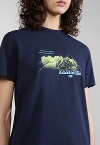 Tumma sininen t-paita, joka on valmistettu puuvillasta. T-paidassa on vaaleanvihreä vuoristografiikka ja valkoinen teksti, sekä logo ja viesti tulostettuna alle.