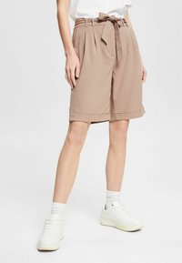 Shorts de corte ajustado en marrón claro con cintura alta, pliegues y un cinturón de tela. Presenta dobladillos enrollados y una textura suave. Llevados con zapatillas blancas.