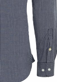Manche longue de chemise bleu marine avec des rayures verticales blanches à pois et un poignet boutonné, montrant la texture du tissu et les détails du motif.