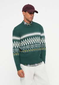 Green knitted sweater featuring a white diamond pattern, with yellow and black accents. Model wears a plaid shirt underneath and a burgundy cap.