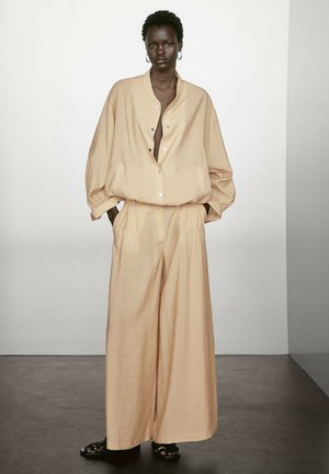 Beige oversized Button-up-Hemd mit fallenden Schultern, kombiniert mit weit geschnittenen Hosen. Eine glatte Textur, ein minimalistisches Design und dunkle Sandalen runden den Look ab.