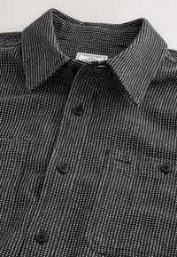 Camisa en blanco y negro con patrón, cuello abotonado, bolsillo en el pecho, tela texturizada y botones negros.