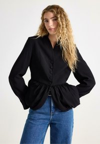 Sort struktureret jakke med peplum-høm, knaplukning og lange ærmer, parret med blå denimjeans. Enkel, moderne design.