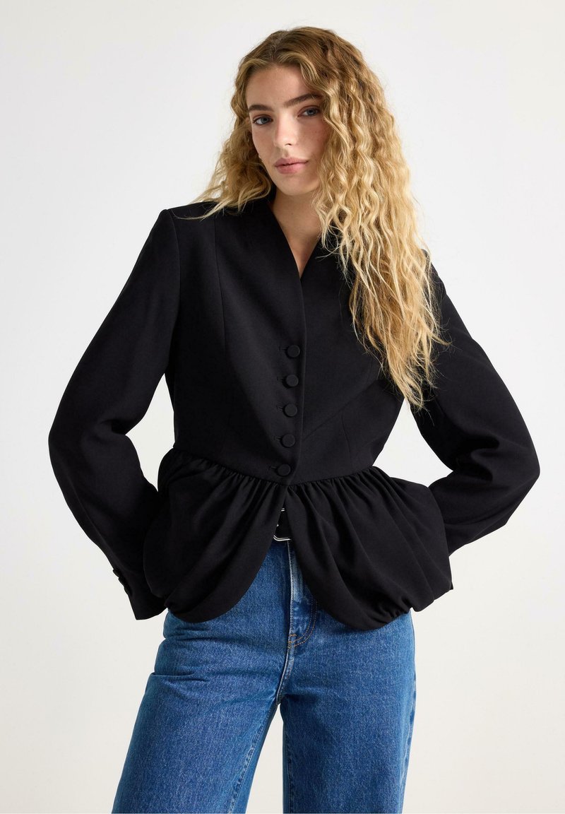 Sort struktureret jakke med peplum-høm, knaplukning og lange ærmer, parret med blå denimjeans. Enkel, moderne design.