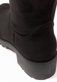 Sort ruskindsankelstøvle med en blokhælhæl, tekstureret overflade og lagdelt design. Gummisål med riflet mønster for bedre traction.