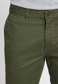 Olivově zelené bavlněné kalhoty s hladkým povrchem, poutky na opasek a knoflíkovým zapínáním v pase. Obsahují boční kapsu.