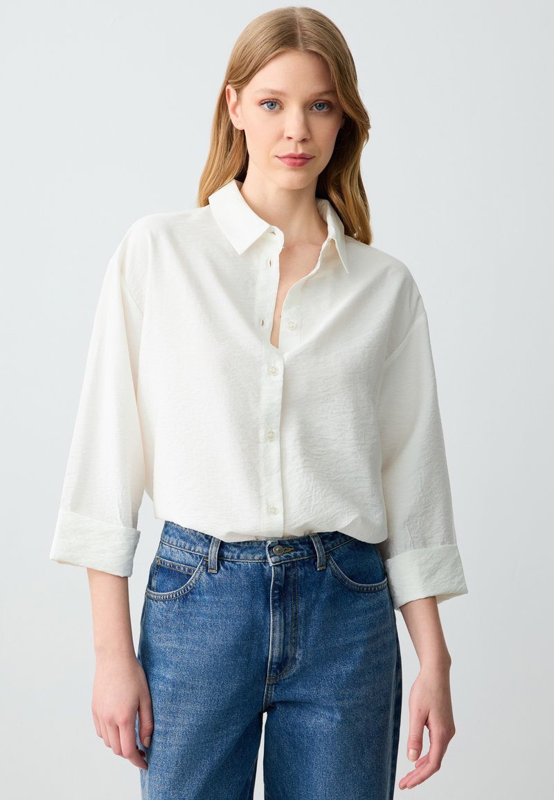 Witte button-up shirt met een textuurafwerking, opgerolde mouwen en een kraag, gecombineerd met hooggegorte blauwe denim jeans.