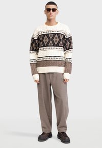 Mand står iført solbriller, mønstret beige og brun striktrøje, løse taupe bukser og mørkebrune sko på hvid baggrund.