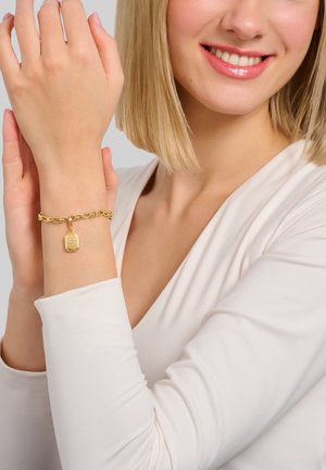 Donna con maglietta bianca a maniche lunghe sorridente, che mostra un bracciale a catena d'oro con ciondolo rettangolare al polso sinistro.