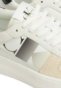 Sneaker bianca con tomaia in pelle liscia, caratterizzata da un grande logo "CK" in gradiente grigio e un accento in suede beige sui lati. Suola in gomma testurizzata.