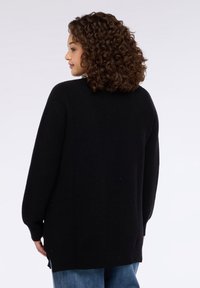Maglione nero a maniche lunghe con vestibilità rilassata, trama a maglia, orlo arrotondato e spacchi laterali, abbinato a jeans azzurri.