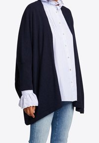 Námořnický kardigan s volně spadenými rameny, otevřeným předním dílem a texturovanou látkou. Pod ním světle modrá košile s proužky a volánovými manžetami.