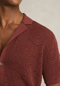 Polo tricotat în roșu închis, cu un model hexagonal și detalii de dantelă deschisă. Dispune de un guler și închidere cu buton.