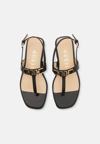Mustat thong-sandaalit, joissa on kultaiset yksityiskohdat, nahkaremmit ja neliön muotoinen kärki. Beige pohja, jossa on "GUESS" -brändäys.