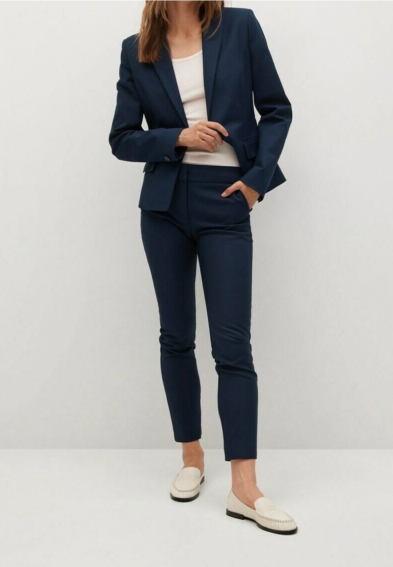 Navy modrý přizpůsobený blazer s odpovídajícími slim kalhotami, s hladkou texturou. Nosený přes světložlutý top, doplněný krémovými mokasíny.