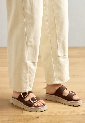 Bruine suède sandalen met twee brede banden, elk met een vierkante metalen gesp, op een dikke beige rubberen zool. Draagt met lichtgekleurde broek.