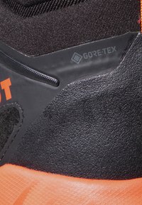 Scarponcino da trekking nero e arancione con materiale in gomma testurizzato, dotato di logo GORE-TEX, colletto imbottito e design della suola robusto.