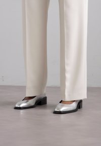 Hõbedased metallik blokknõeltega jalanõud ruudukujulise varbaga, mida on kombineeritud koorevärvi kohandatud pükstega. Sile tekstuur ja sujuv viimistlus.