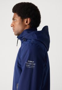 Veste imperméable navy avec un col haut, une capuche réglable et des matériaux écologiques. Comprend des détails de logo et de texte sur la manche.