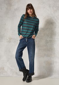 Grün-graues gestreiftes Langarmshirt, kombiniert mit lockeren blauen Jeans mit Knopfverschluss und aufgerollten Säumen, ergänzt durch schwarze Stiefel.