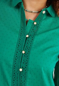 Chemise verte boutonnée en tissu doux, arborant un panneau en dentelle sur le devant, avec des boutons nacrés et des motifs texturés.