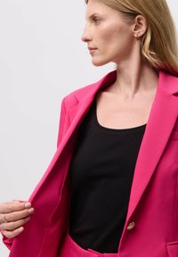 Fuksija sako sa strukturiranim ramenima, reverima i jednim zlatnim dugmetom, nosi se preko crnog topa. Glatka tekstura tkanine.