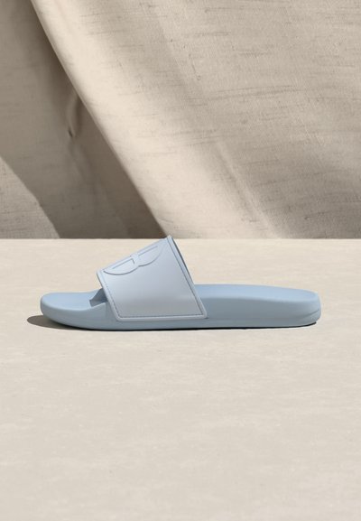 Sandale à glissière bleu clair avec une large bride et un logo discrètement embossé, posée sur une surface beige lisse avec un fond en tissu.