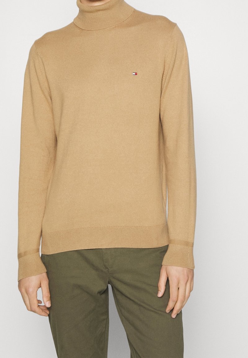 Pull à col roulé beige avec manches longues et un petit logo rouge-blanc-bleu sur la poitrine, porté avec un pantalon vert olive.