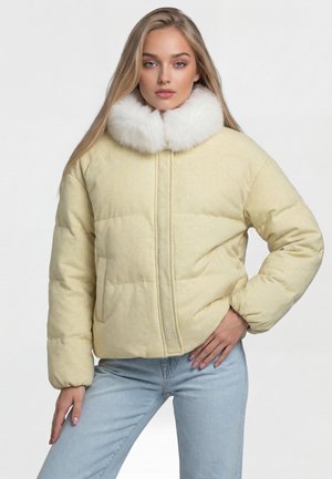 Junge Frau trägt eine hellgelbe Steppjacke mit weißem Pelzkragen, posiert mit einer Hand in der Hüfte und der anderen entspannt an der Seite.