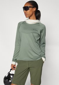 Maglietta verde a maniche lunghe con scollatura bianca, con logo. Abbinata a pantaloni corti oliva. Il modello tiene un casco nero. Indossa occhiali da sole.