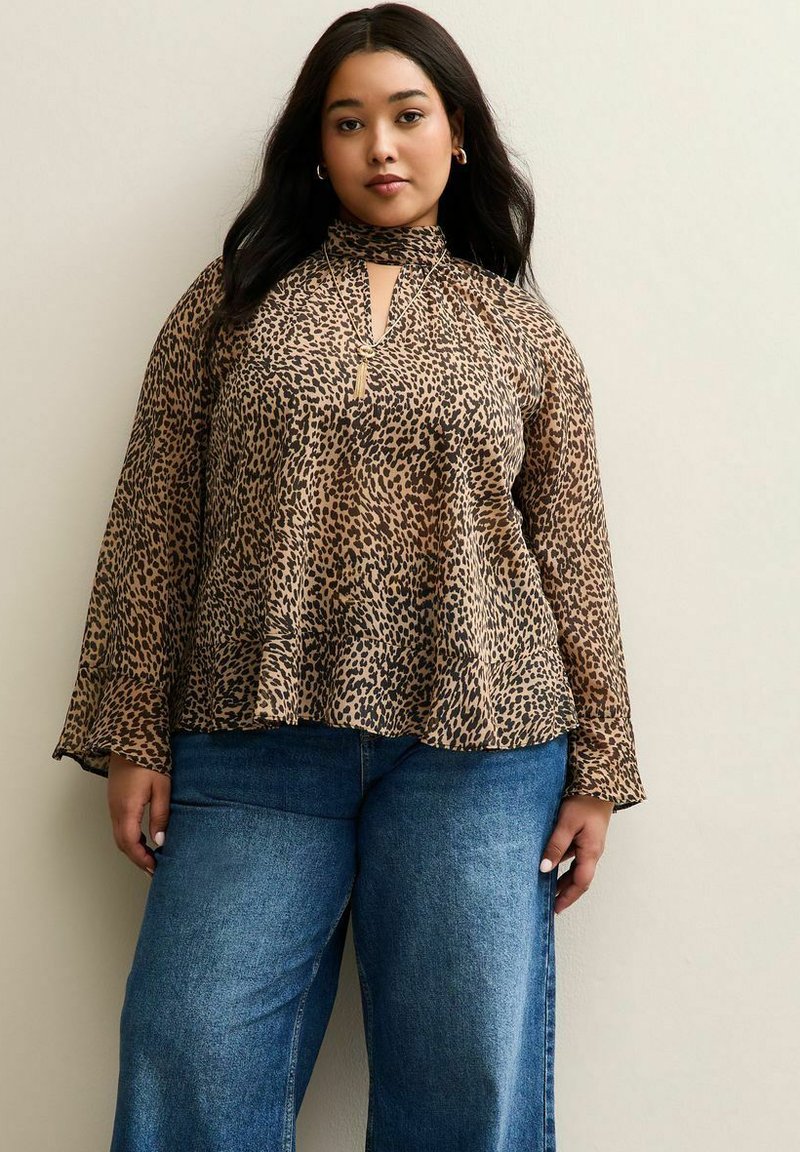 Kobieta stojąca przy jasnej ścianie, ubrana w brązową bluzkę w leoparda z długim rękawem oraz niebieskie szerokie dżinsy, nosząca złote kolczyki w kształcie kół oraz naszyjnik.