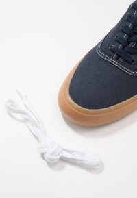 Sneaker in tela blu marina con suola in gomma color caramello e cuciture bianche. Presenta un'intricate stringatura e un laccio bianco annodato nelle vicinanze.