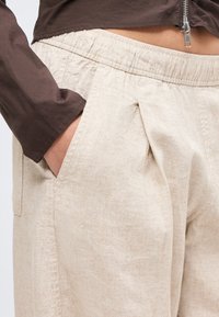 Χέρι στην τσέπη μπεζ παντελονιού με υφή και ελαστική μέση, σε συνδυασμό με σκούρο καφέ μακρυμάνικη μπλούζα με λεπτομέρεια φερμουάρ.