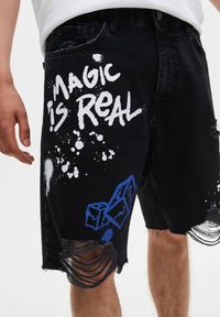 Shorts en denim noir avec un ourlet effiloché, arborant un texte en spray blanc "LA MAGIE EST RÉELLE" et des accents graphiques bleus.
