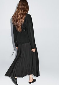 Černý pletený svetr s volným střihem, doplněný o splývavou černou plisovanou sukni a černé slip-on boty. Dlouhé vlnité vlasy jsou viditelné.