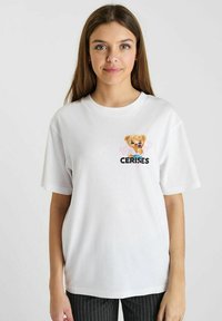 T-shirt blanc en coton à manches courtes avec un graphique d'ours en peluche tenant un cornet de glace, accompagné d'accents en forme de cœur rose et du texte "CERISES."
