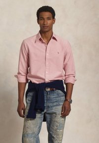 Ljusrosa långärmad skjorta med knapp-down krage, bärs över en marinblå tröja knuten vid midjan, i kombination med slitna blå jeans.