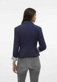 Marinblå blazer med en figursydd design och snedskurna slag, med randiga manschetter. Matcher med ljusgrå jeans. Slät tygtextur.