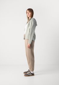 Šviesiai žalia zip-up gobtuvu striukė, vilkima ant baltos marškinėlių. Smėlio spalvos džinsai su elastinėmis rankovėmis, derinami su pilkais sportbačiais ir baltais kojiniais.
