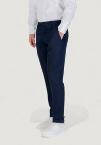 Homme portant un pantalon slim-fit bleu marine, une chemise blanche à manches longues et des baskets blanches, se tenant avec une main dans la poche.